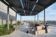 Students study and relax on a rooftop terrace featuring wooden benches and tables shaded by a solar panel canopy, with a campus and city skyline in the background.