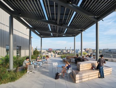 Students study and relax on a rooftop terrace featuring wooden benches and tables shaded by a solar panel canopy, with a campus and city skyline in the background.