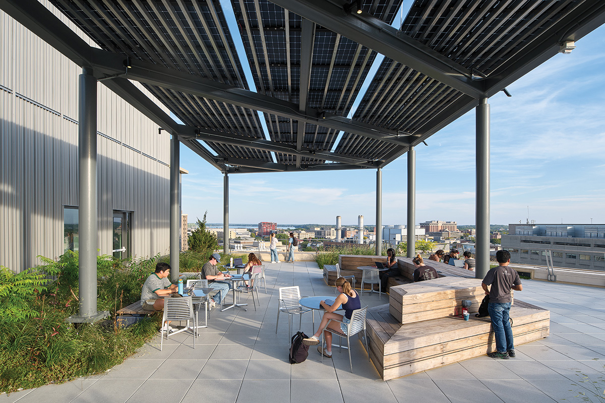Students study and relax on a rooftop terrace featuring wooden benches and tables shaded by a solar panel canopy, with a campus and city skyline in the background.