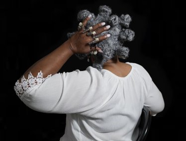 Seen from behind against a black background, a person wearing a white lace-trimmed top rests a hand adorned with multiple pearl rings on a wig of gray hair styled in Bantu knots.