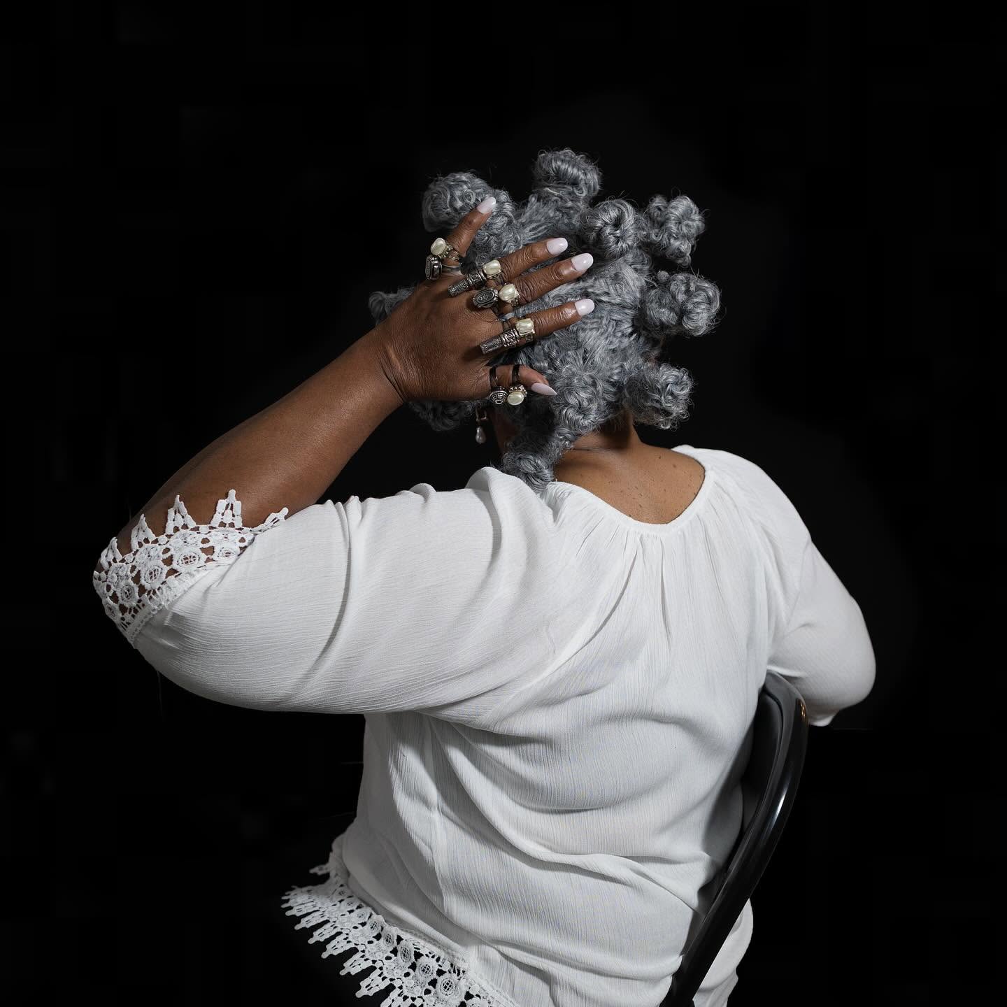 Seen from behind against a black background, a person wearing a white lace-trimmed top rests a hand adorned with multiple pearl rings on a wig of gray hair styled in Bantu knots.