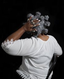 Seen from behind against a black background, a person wearing a white lace-trimmed top rests a hand adorned with multiple pearl rings on a wig of gray hair styled in Bantu knots.