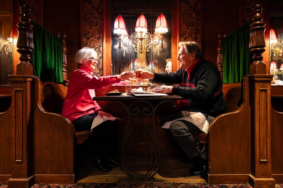 A couple toasts with drinks while seated across from each other in a cozy, wooden dining booth under warm restaurant lighting.