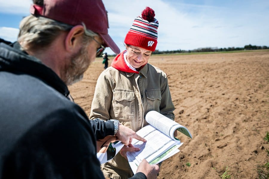A woman wearing a red Wisconsin stocking hat and tan jacket reviews a document with a colleague in a tilled agricultural field.