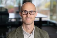 Portrait of Simon Goldberg wearing black-framed glasses and an olive green blazer.