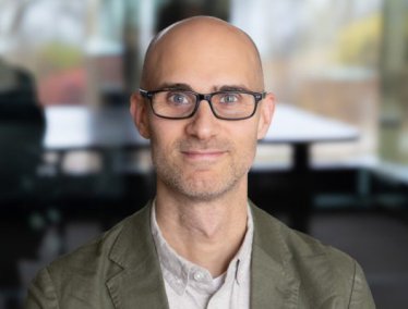 Portrait of Simon Goldberg wearing black-framed glasses and an olive green blazer.