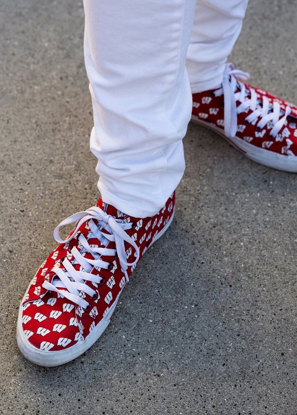 A close-up view of someone wearing white pants and red canvas sneakers patterned with white Wisconsin Badgers 'W' logos.