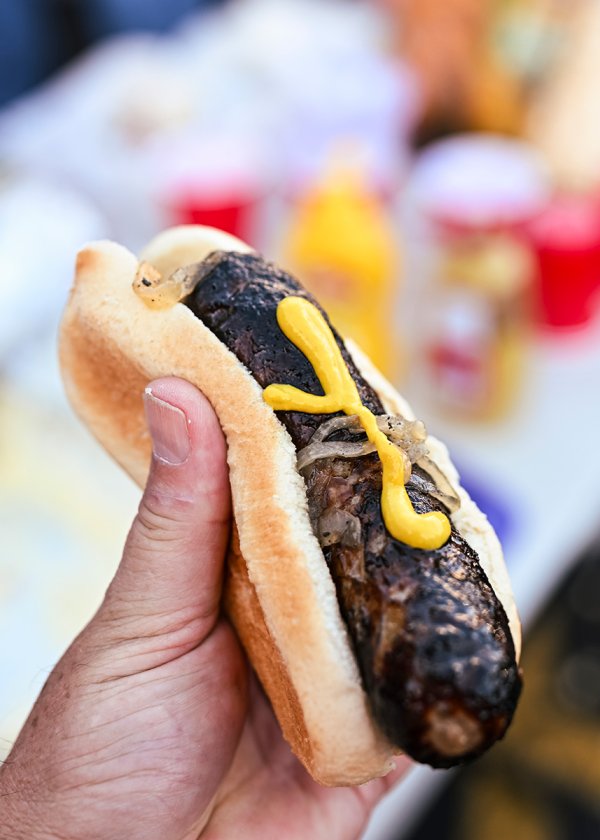 A hand holds a grilled bratwurst in a white bun topped with grilled onions and yellow mustard.