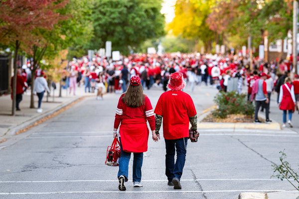 A couple wearing red Wisconsin Badgers clothing holds hands while walking down State Street, filled with Badger fans, in autumn.