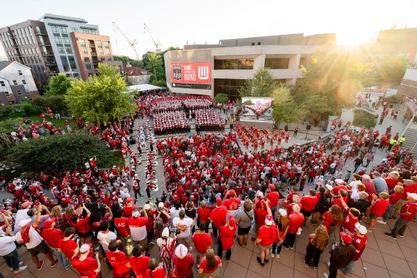 A high-angle view shows a large crowd of fans in red gathered in a plaza outside Union South watching the marching band perform.