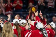 Members of Wisconsin's women's hockey team wearing red jerseys celebrate their victory by hoisting an NCAA championship trophy overhead.