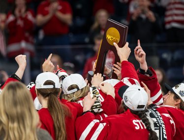 Members of Wisconsin's women's hockey team wearing red jerseys celebrate their victory by hoisting an NCAA championship trophy overhead.