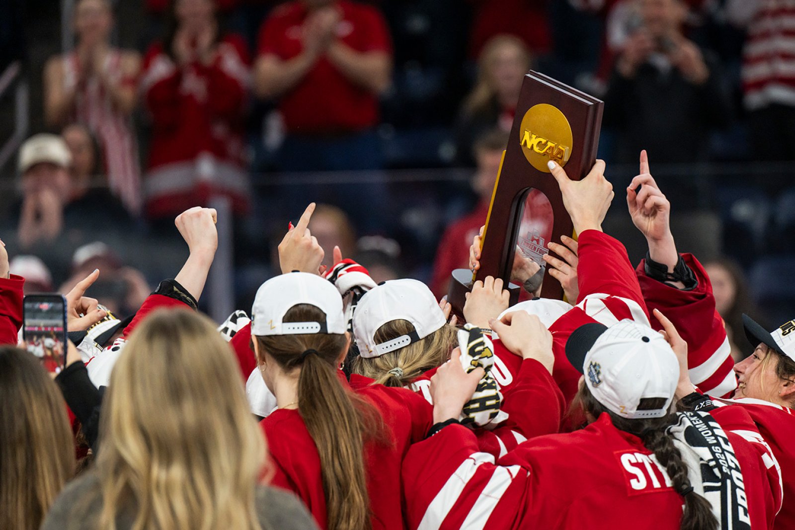 Members of Wisconsin's women's hockey team wearing red jerseys celebrate their victory by hoisting an NCAA championship trophy overhead.