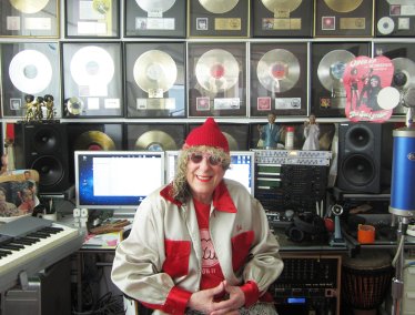 Songwriter Allee Willis sits smiling in a music studio surrounded by audio equipment and a wall filled with framed gold and platinum record awards.