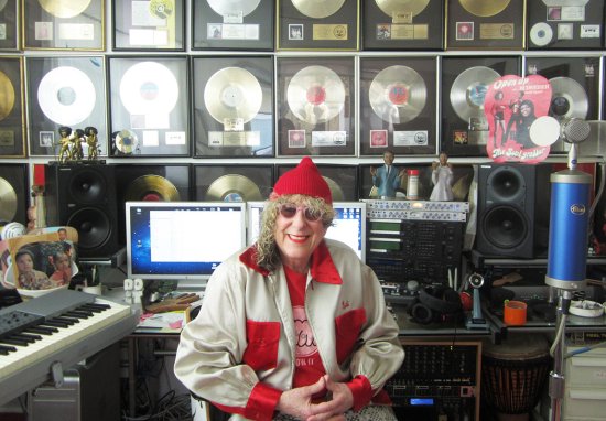 Songwriter Allee Willis sits smiling in a music studio surrounded by audio equipment and a wall filled with framed gold and platinum record awards.