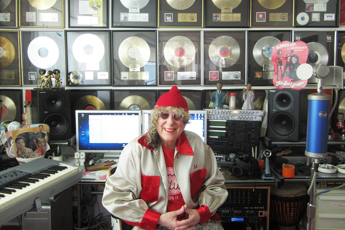 Songwriter Allee Willis sits smiling in a music studio surrounded by audio equipment and a wall filled with framed gold and platinum record awards.